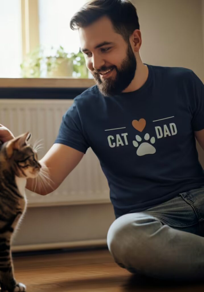 Why Cat Owners Love Graphic Tees 2 A man wearing a 'Cat Dad' shirt playing with his tabby cat on the floor.