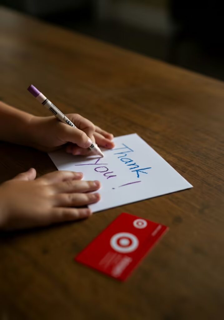 A close-up of a child's hands writing a thank you card for their teacher, with a gift card nearby.